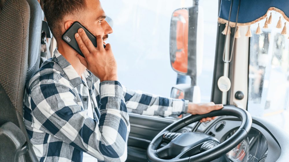 Talking by phone. Young truck driver is with his vehicle at daytime.
